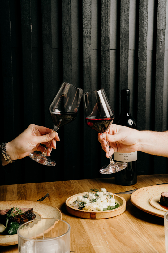Two diners clinking glasses of red wine at Peonee Restaurant Canberra during dinner.