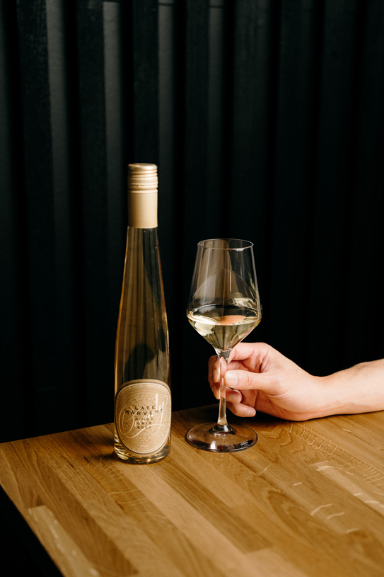 Hand holding a glass of white wine beside a bottle on a wooden table at Peonee Restaurant Canberra.